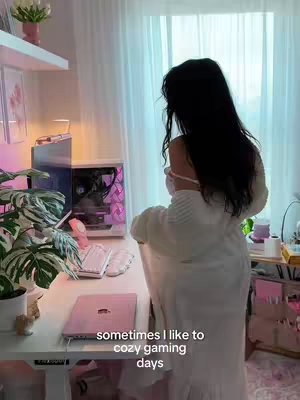 A woman with long black hair is standing in front of a desk with a monitor and a laptop, likely in her room. She is wearing a white outfit and is looking away from the camera towards a window. The lighting suggests a sunny indoor setting. Text overlayed on the image reads: "sometimes i like to cozy gaming days"