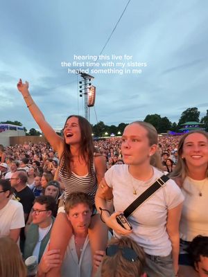 A crowd at an outdoor concert with three young women. The woman in the center is raised on someone's shoulders, with an arm raised in the air. Text overlay at the top.