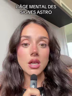 A woman with long, dark hair and makeup faces the camera with an open mouth and a microphone held close. Above her, a text overlay reads, 'L'AGE MENTAL DES SIGNES ASTRO'. The background is softly out of focus.