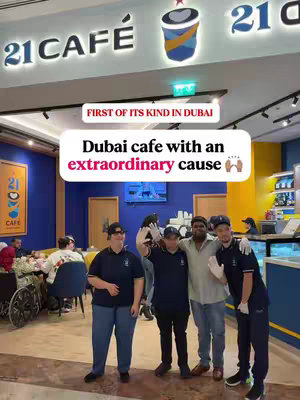 The image shows the interior of "21 Cafe" with several people. Text overlays indicate that it's the first of its kind in Dubai, describing it as a cafe with an extraordinary cause. The cafe staff and customers are in the background, some wearing black polo shirts. The overall setting seems supportive and happy.