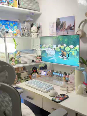 A well-lit room with a desk setup, featuring a large monitor displaying an illustration of a cat on a boat. The desk is organized with a keyboard, mouse, and various decorative items like figurines and toys. Shelves hold additional collectibles and accessories. The overall aesthetic suggests a cozy, personalized workspace.