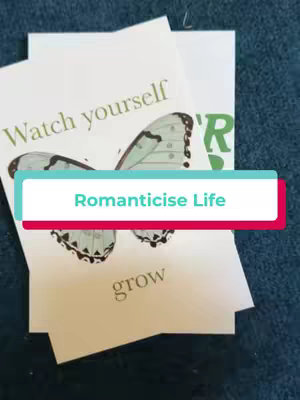 The cover image shows a fan of printed photographs, with the central focus on a rectangular overlay that reads "Romanticise Life." The background features a dark blue surface which seems to be the surface on which the photo-prints are set. Layered behind it, some printed photos can be partially seen. These feature illustrations, written content, and are printed on a white base. One photo shows a butterfly and the text saying "Watch yourself", and "grow". The overlay in the middle is a red rectangle where the text "Romanticise Life" is in white font. The image and the overlay are in soft lighting