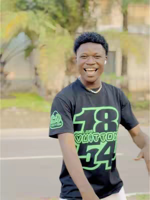 A young man with a dark complexion, wearing a black t-shirt with a green graphic design, is smiling widely with his mouth open. He has short, dark, curly hair and is wearing a single earbud. He has a thin necklace and is standing outside on a sunny day, with greenery in the background. The composition is a medium shot.
