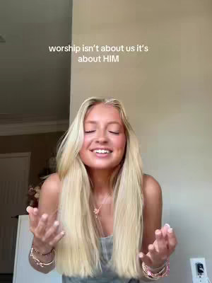 A woman with long blonde hair is shown smiling with her hands held up, palms facing the camera. The background is a light, neutral color and the text overlay reads "worship isn't about us it's about HIM".