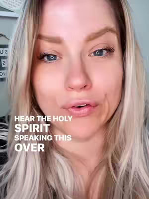 A close-up shot features a fair-skinned woman with long, light blonde hair. Her blue eyes are framed by thick lashes, and she is looking slightly downwards. In the lower portion of the frame, text reading "HEAR THE HOLY SPIRIT SPEAKING THIS OVER" is visible.