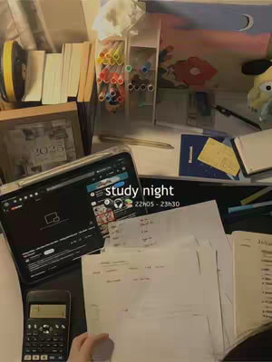 A desk setup with an iPad, books, and stationery items. The scene is lit with warm artificial light, and there are papers spread out, suggesting a study environment. The color palette is muted, with whites, browns, and touches of color from the stationery.