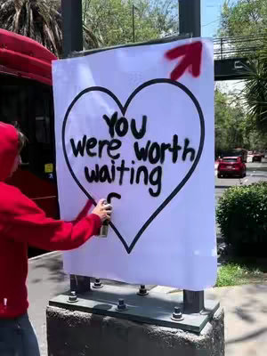 A person is spray painting the phrase "YOU were worth waiting" inside a large outline of a heart on a white sheet. An arrow points upwards next to the heart. The person, wearing a red hoodie, is holding a spray paint can. A red bus and a street with traffic are in the background.