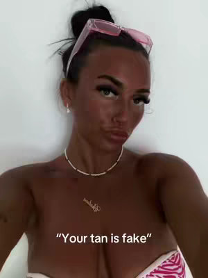 A woman with dark hair pulled back in a bun, wearing pink sunglasses on her head, a white necklace, and a patterned bikini top. She has a deep tan, visible makeup, and is making a slight face.