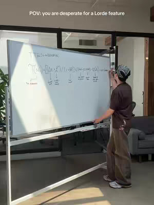 A person is standing next to a whiteboard, writing on it. The whiteboard has equations and arrows drawn on it. The person is wearing a bandana, a brown shirt, and khaki pants. The lighting is bright, and the background shows an office setting.