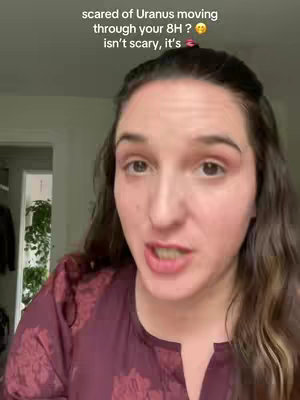 A woman with long brown hair is centered in a medium shot. She's wearing a maroon patterned blouse and speaking towards to camera. Above her is text that reads, "scared of Uranus moving through your 8H?isn't scary, it's". The background includes glimpses of an indoor setting.