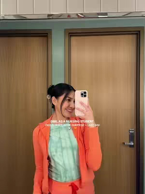 A woman in an orange jacket and green top is taking a mirror selfie in front of two closed wooden doors. She is holding a phone up to her face. Text overlays the image promoting a product and referencing nursing.