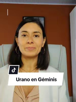 A woman with dark hair and hoop earrings is centered in the image. She is wearing a light colored blazer over a top. Below her is a white rectangle with black text on it and social media icons. The background is composed of a warm orange wall and a chair.