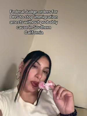 A woman with dark hair and hoop earrings is shown in a close-up shot, holding a pink object near her lips. Text in the background reads, "Federal Judge orders for DHS to stop immigration arrests without probably cause in Southern California."