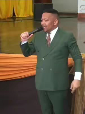 A man in a green suit is holding a microphone and singing, with a backdrop of drapes.