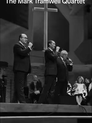 A black and white photo of a group of men singing on a stage. A large cross is visible behind them, lit up in the background. The men are all wearing suits and are holding microphones.