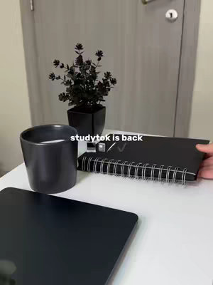 A top-down shot of a workspace featuring a white table with a black notebook, a black ceramic cup, and a black tablet. A small potted plant in a black pot is positioned to the side against a gray door. The text "studytok is back" is overlaid.