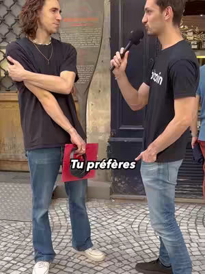 Two men are standing on cobblestone and talking. The man on the left is wearing black T-shirt and holding a red object. The man on the right is wearing a black T-shirt with white text, and holding a microphone. Text overlay indicates the title of the video.