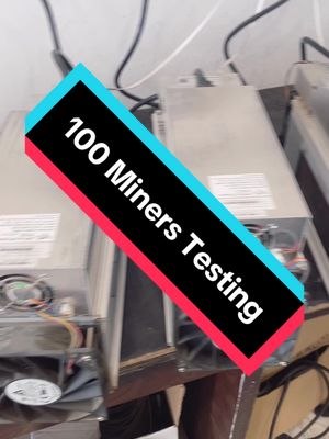 The image shows a close-up of several cryptocurrency mining machines lined up, with a diagonally placed text overlay highlighting the testing of 100 units. The background is blurry, and the color scheme is dominated by the metallic gray of the machines, contrasted by the black and red of the overlay.