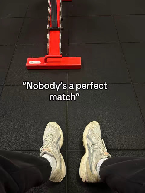 A first-person view of a person's feet in light-colored sneakers on a black gym floor. Above, a red weight rack is visible, and white text overlays with a quote.