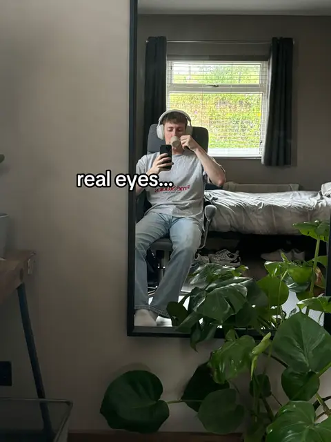 A man in a reflective shot in a mirror, sitting in a chair with headphones. The mirror captures a view of a room with a window and bed in the background. A text overlay reads "real eyes..."