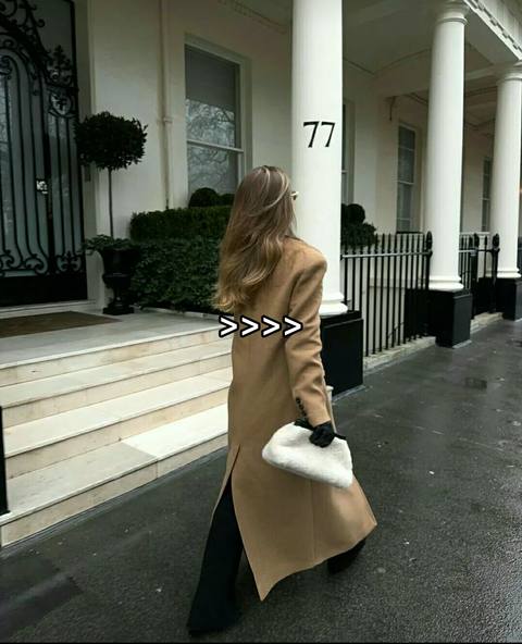 A woman in a long, camel-colored coat walks away from the camera on a wet street in front of a classical building. She holds a white fluffy clutch. The image is slightly desaturated. The word "77" is on the building behind her, and the image has a series of arrows as a text overlay.