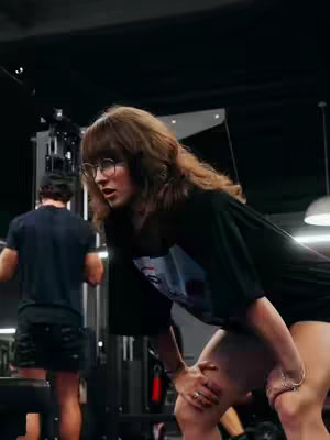 A woman in workout attire leans over, hands on knees, in a gym setting. She has brown hair and glasses. Another person is visible in the background.
