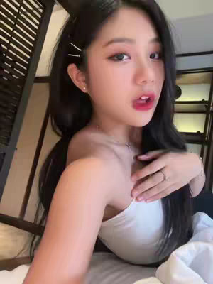 A selfie-style shot of a woman in a white tube top, reclining on a bed. She has long black hair styled with hair clips. She has red lipstick, she gazes directly at the camera with a slightly open mouth, with a hand reaching towards her chest.