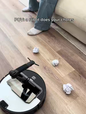 A robot vacuum cleaner on a wooden floor with crumpled paper scattered around it. The POV shot shows the vacuum approaching the viewer, next to a person seated on a beige couch. Text overlay reads 'POV: a robot does your chores'.