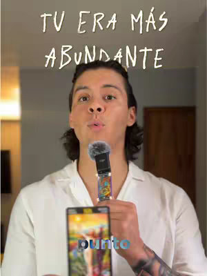 The cover image shows a man with dark hair and a white shirt holding a microphone. Above his head is the text 'TU ERA MÁS ABUNDANTE' in a handwritten style. He holds a smartphone displaying the word 'punto' in a colorful image. The background suggests an indoor setting with neutral tones.