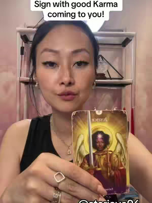 A woman with long dark hair and fair skin holds up a tarot card. She is positioned in front of a white shelf, and the background has a soft, warm light. The tarot card has an illustrative style card with an angel-like figure. The woman looks directly at the viewer with a gentle expression, her lips slightly parted. Behind her, there is a soft, warm light.