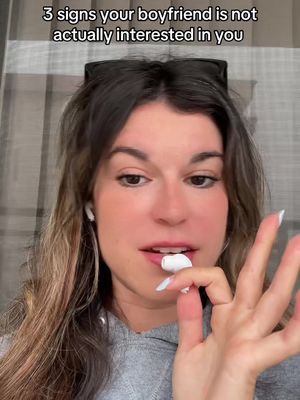 A woman with long brown hair is looking at the camera. She is in a grey hoodie with earbuds in and is holding one white earbud between her teeth with her index finger near her lips. Text at the top says '3 signs your boyfriend is not actually interested in you'.