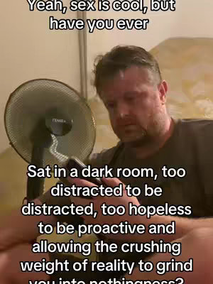 A man is sitting in a room with a fan running, looking down with a somber expression. Text overlays the image.
