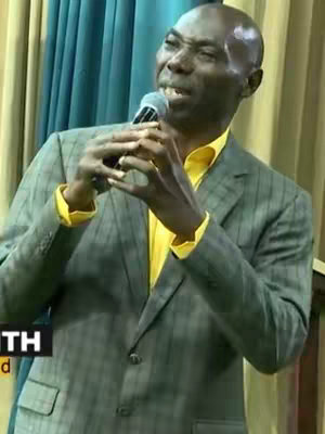A man in a gray suit and yellow shirt speaks into a microphone, with a supportive background of curtains. He appears to be delivering a speech or a sermon.