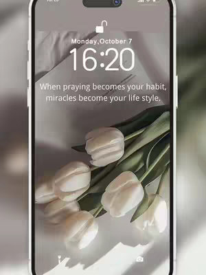 The image, framed in a vertical orientation as if a mobile phone display, features a close-up of white tulip blooms arranged on a light, off-white surface. Above the floral arrangement, within the upper portion of a smartphone display, the time "16:20" is displayed in a clean, white font. The phone's lock screen displays the date, "Monday, October 7", and features a quote that reads, "When praying becomes your habit, miracles become your life style." The overall composition is soft, the lighting is gentle, and the style is modern and serene. The phone itself appears to be white or a light color.