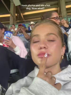 A selfie-style shot taken in what appears to be a stadium setting with a woman as the central figure. The woman is fair-skinned with blonde hair, wearing a light gray hoodie and multiple rings on her fingers. She has her index finger playfully raised toward her lips, with her eyes closed. The background includes blur of other spectators and stadium seating. The lighting indicates that it is a bright day. The text overlay is positioned at the top of the frame.