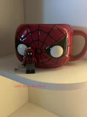 The cover image shows a Spiderman-themed mug placed on a white shelf. Beside the mug is a Lego Miles Morales keychain. The mug is red with a black spiderweb design on its front and large white circles where the eyes would be. The Lego keychain is a miniature figure of the Miles Morales Spiderman character. The background appears to be an interior setting. The overall color palette is dominated by red, white, and black, creating a fun, playful composition.