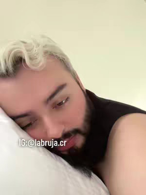 A man with white hair and a beard lies on a white pillow, looking down with a thoughtful expression. The image focuses on his face, and the lighting appears soft.  Social media information is in the corner.