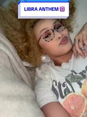 A woman lies on a bed, head on a pillow, looking toward the viewer. She has curly blond hair and wears glasses. A text box with the words "Libra Anthem" is at the top of the image. The woman looks directly at the camera with a neutral expression.  She is wearing a t-shirt with a design on it as well.