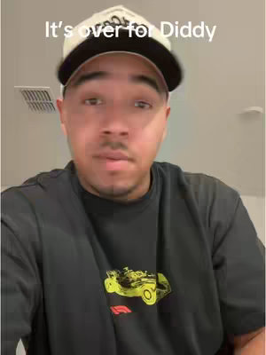 A man wearing a white and black baseball cap and a black t-shirt with a yellow design is looking at the camera. Text overlay reads "It's over for Diddy".