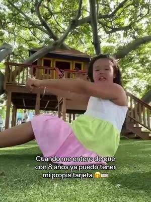 A young girl sits on the grass with a treehouse in the background. She wears a white top, a pink skirt and a green skirt over it. The child is making a gesture with her arms as if wanting to show something. The background is a field with greenery.