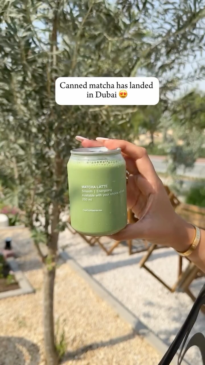 A hand holding a canned matcha drink against a blurred background of greenery and sunlight. The can is green, matching the drink inside, and features white text. A smaller white rectangular text box at the top displays the hook text, which includes the phrase 'Canned matcha has landed in Dubai' with a heart-eyes emoji.
