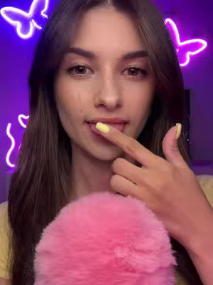 A young woman with long brown hair is shown in a close-up shot. She has fair skin and is looking at the camera with a soft expression. Her index finger is touching her lip, and she has yellow nail polish on her nails. Behind her, in the purple background, are neon butterfly lights.