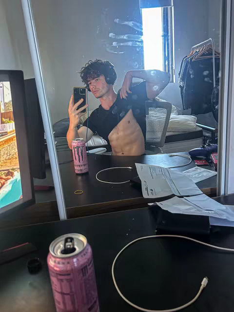 A man takes a selfie in front of a mirror at home. He is wearing headphones and a black shirt, which is partially lifted to expose his abs. He is holding a phone in his hand taking the picture. Around him are objects like a pink Monster energy drink, a monitor, and some papers, all in a messy room.