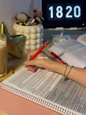 A person's hand, adorned with a bracelet, is shown writing on a notebook with a red pen. The notebook is open on a desk next to an open book. In the background, a digital clock displays the time and a decorative container holds makeup brushes. A cup of coffee is visible on the left, with a star ornament next to the cup. The overall theme suggests a study environment.