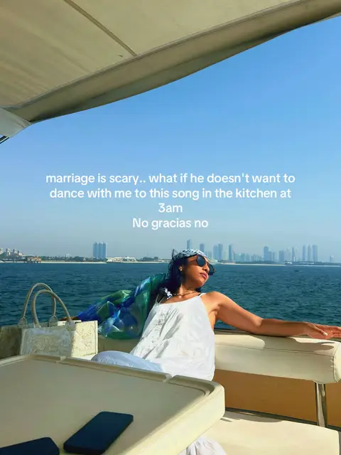 A woman reclines on a white yacht seating area under a shaded covering, facing right with the sea and city skyline behind. She wears sunglasses, white clothing, and a patterned headscarf. Text overlay is present.