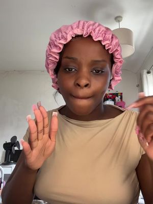 A young Black woman is shown in a close-up shot, appearing to be in her home. She's wearing a pink satin bonnet and a light brown top. The woman is looking at the camera with a slight smirk and has one hand up near her face with her fingers spread, while the other arm is by her side with the pointer and middle finger extended at the camera. The background is slightly blurred but shows a neutral-toned wall with a pendant lamp and a possible piece of artwork.