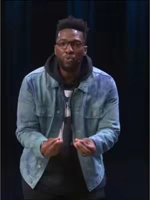 The image features a man in a denim jacket and glasses, standing against a dark background. He has dark skin and short, styled hair. He's wearing a black hoodie underneath the jacket. His hands are up in a gesture. The lighting is focused on the man, highlighting his face and upper body.