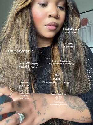 A selfie-style shot featuring a person from the chest up, positioned slightly off-center. The person has long, brown hair with blonde highlights. They wear a black top and multiple rings on their fingers. The person's left arm is visible, also decorated with tattoos. Overlaid text annotations appear across the image, suggesting a fortune-telling or prediction theme.