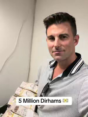 A man in a gray polo shirt stands and smiles, holding a stack of packaged bundles of currency. Behind him are more currency bundles. The background is a plain white wall. Text overlay reading '5 Million Dirhams' with a money bag emoji is at the bottom of the screen.