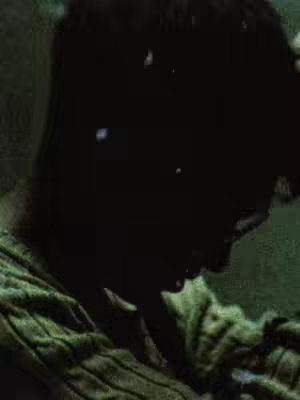 A dark, close-up shot featuring a person's silhouette with a textured fabric visible, possibly a sweater or shirt. The lighting is dim, creating a mood of mystery or introspection.